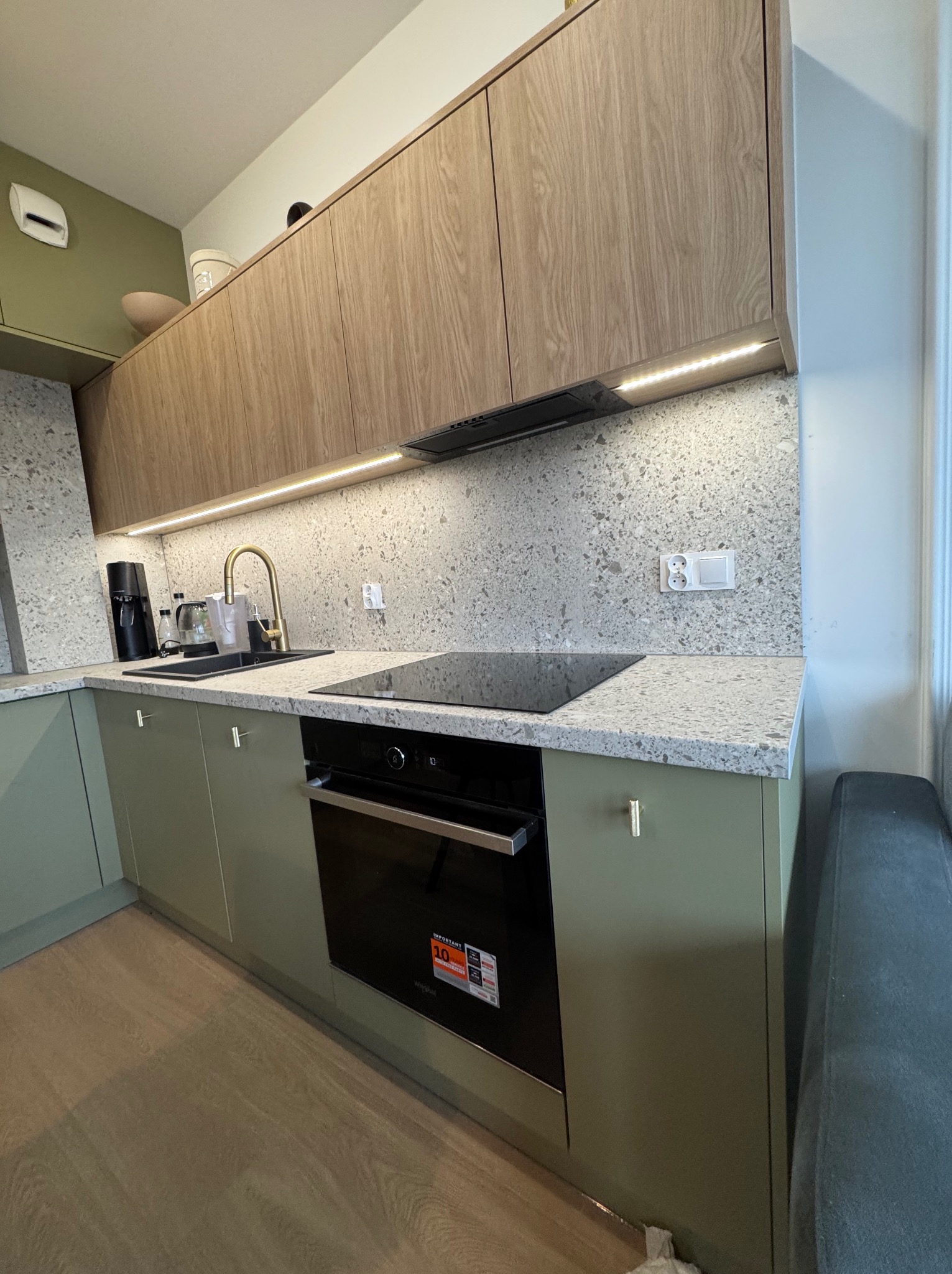 Nowoczesna kuchnia z zielonymi szafkami, drewnianymi szafkami górnymi, blatem z szarego lastryko i złotą armaturą, oświetlona listwą LED pod szafkami.