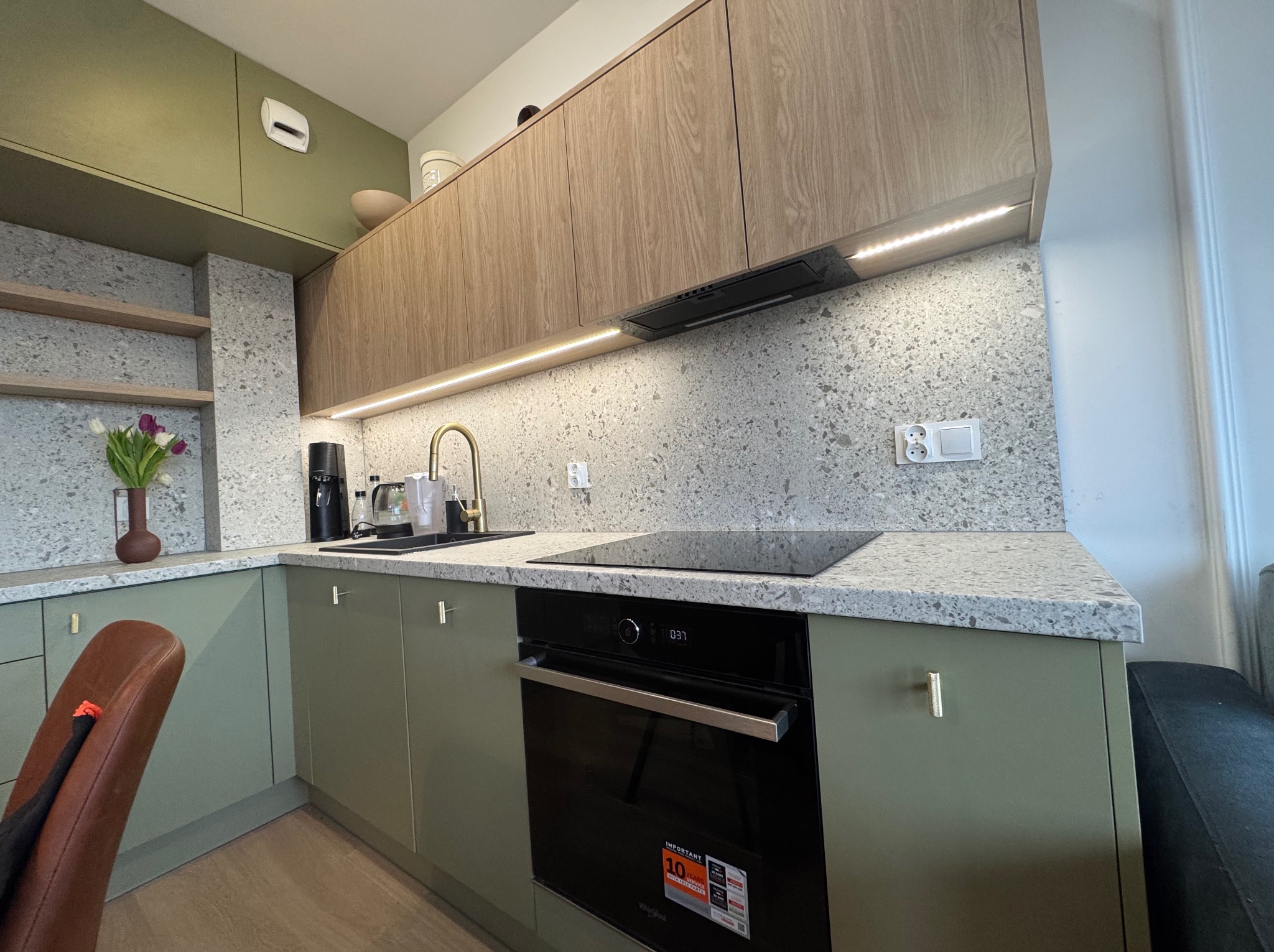 Nowoczesna kuchnia z drewnianymi górnymi szafkami, zielonymi dolnymi szafkami, blatem z szarego lastryko, złotą baterią i czarnym piekarnikiem.