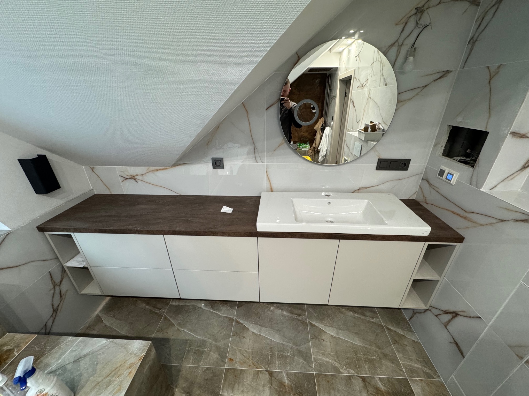 Biała szafka łazienkowa z ciemnobrązowym blatem pod umywalką, zamontowana w łazience na poddaszu z marmurowymi płytkami na ścianach i podłodze; okrągłe lustro odbija osobę robiącą zdjęcie.