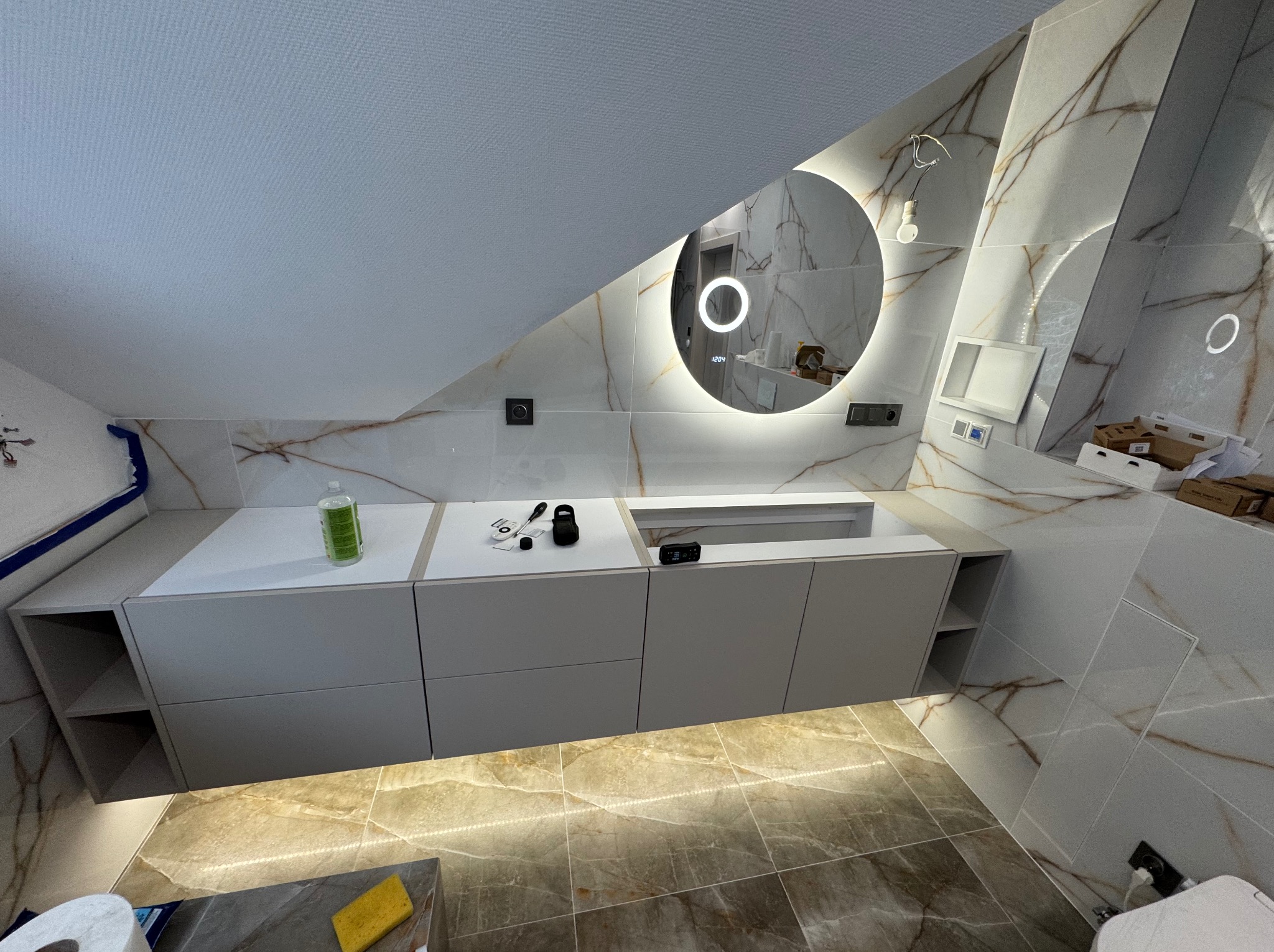 Łazienka na poddaszu z marmurowymi płytkami, okrągłym lustrem z podświetleniem LED i szarą szafką z blatem, widoczne narzędzia i butelka płynu.