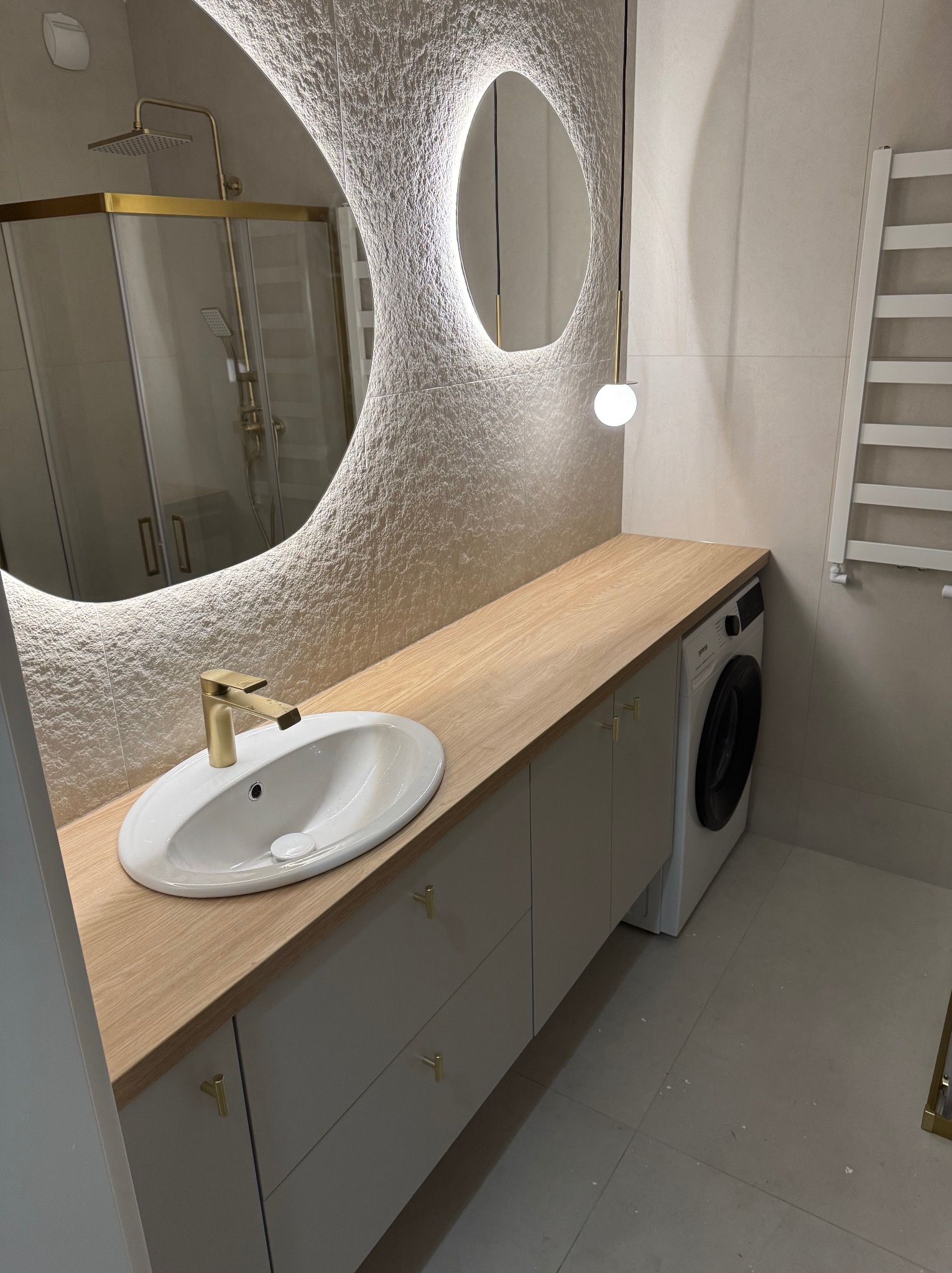 Nowoczesna łazienka z owalnym lustrem podświetlonym LED, drewnianym blatem, białą umywalką z złotą armaturą i szafkami w kolorze złamanej bieli, obok pralka.