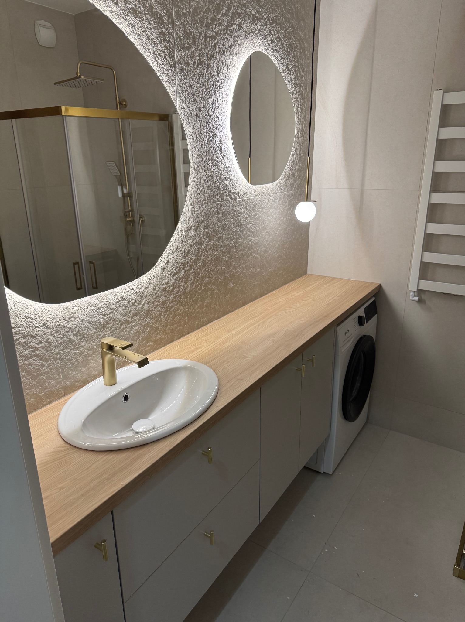 Nowoczesna łazienka z umywalką nablatową, złotą baterią, blatem drewnianym, szafkami w kolorze szarym, pralką i podświetlanym lustrem o nieregularnym kształcie.