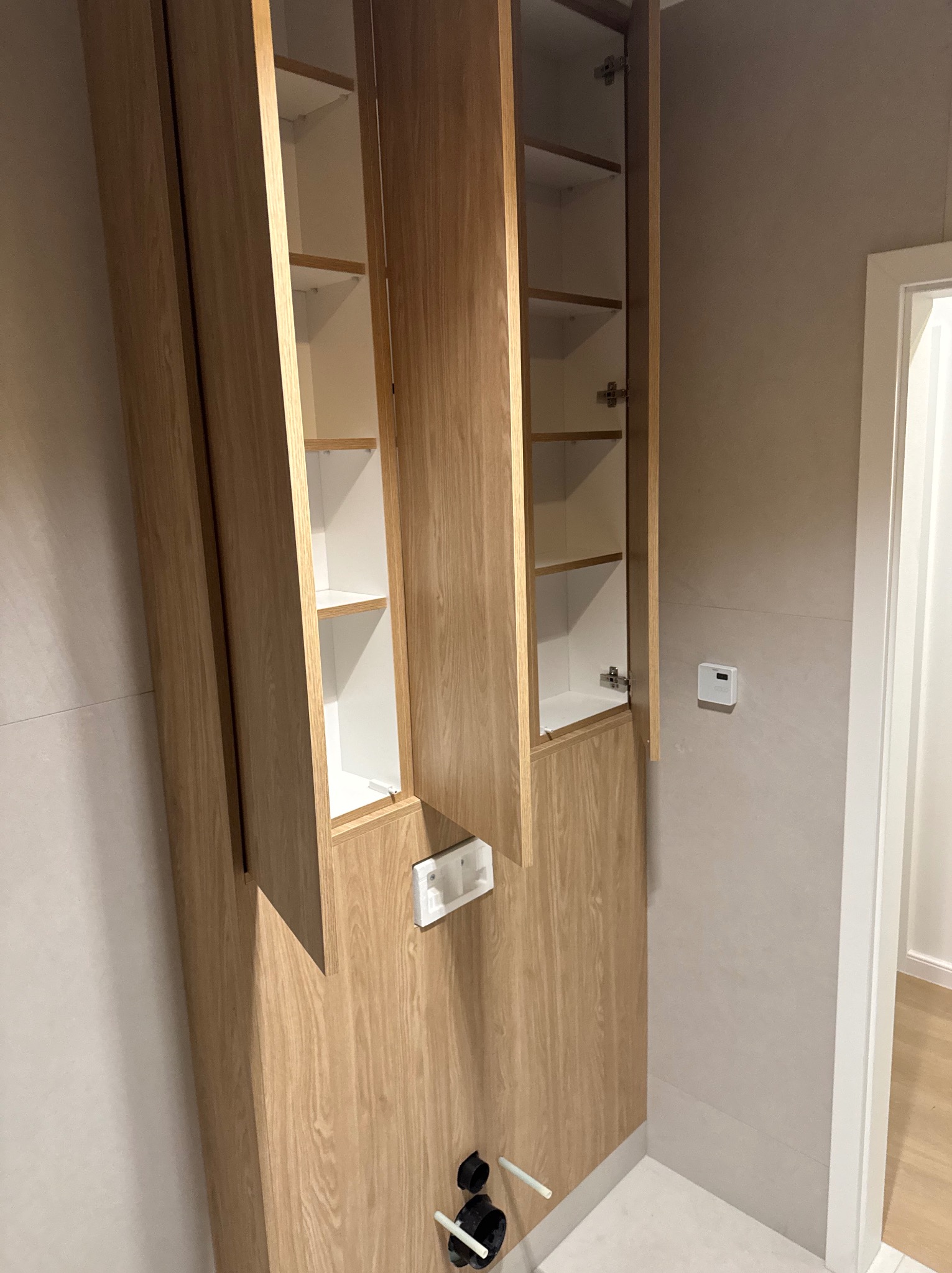 Wysoka, drewniana szafka łazienkowa z otwartymi drzwiczkami, ukazująca białe półki wewnątrz, zamontowana na ścianie z szarą płytką, widoczny fragment instalacji wodnej i biała rama drzwi w tle.