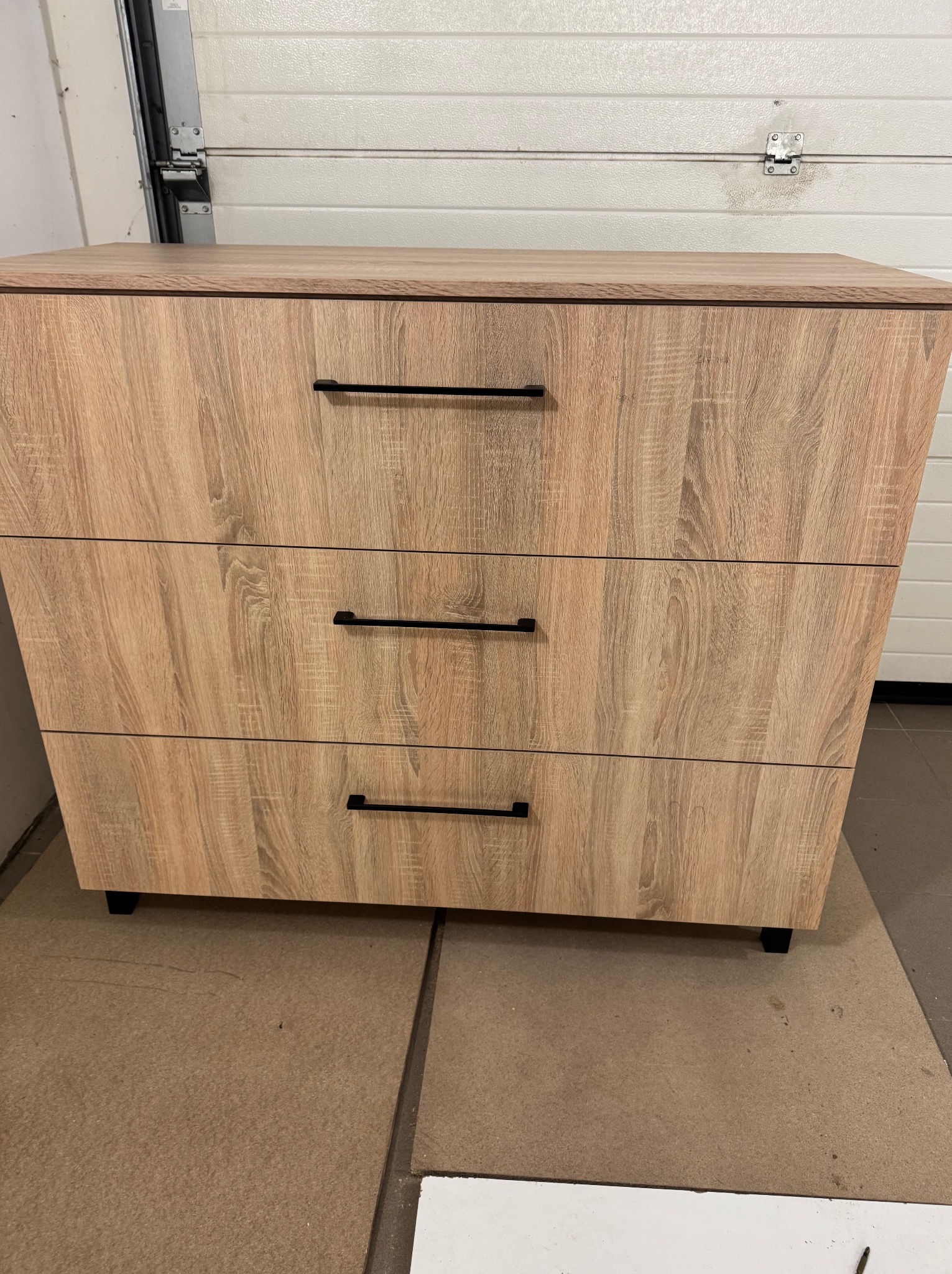 Komoda z trzema szufladami w kolorze jasnego drewna z czarnymi, prostymi uchwytami, wykonana w Iławie, stoi na podłodze wyłożonej płytami pilśniowymi.