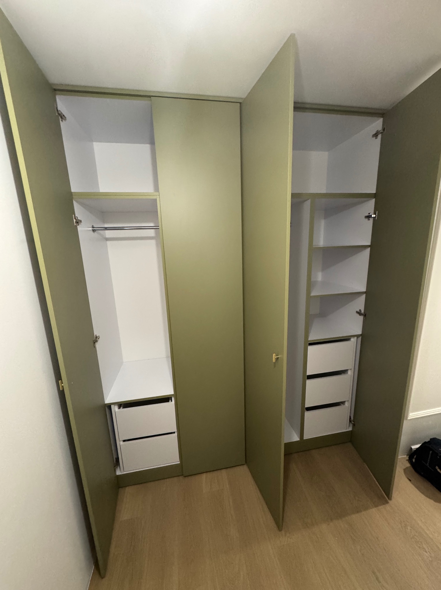 Nowoczesna szafa na wymiar z oliwkowymi frontami, otwarte drzwi ukazują białe wnętrze z szufladami i półkami, widoczne okucia i drewniana podłoga.