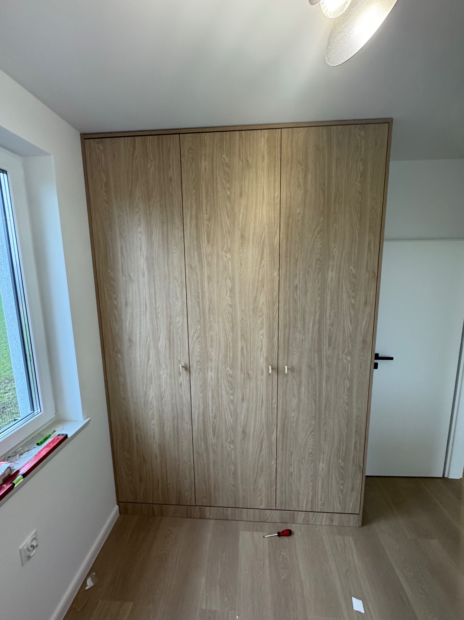 Czterodrzwiowa szafa wnękowa z jasnego drewna z drobnymi, złotymi uchwytami, zamontowana w pokoju z parkietem i białymi ścianami. Na podłodze leży czerwony śrubokręt.