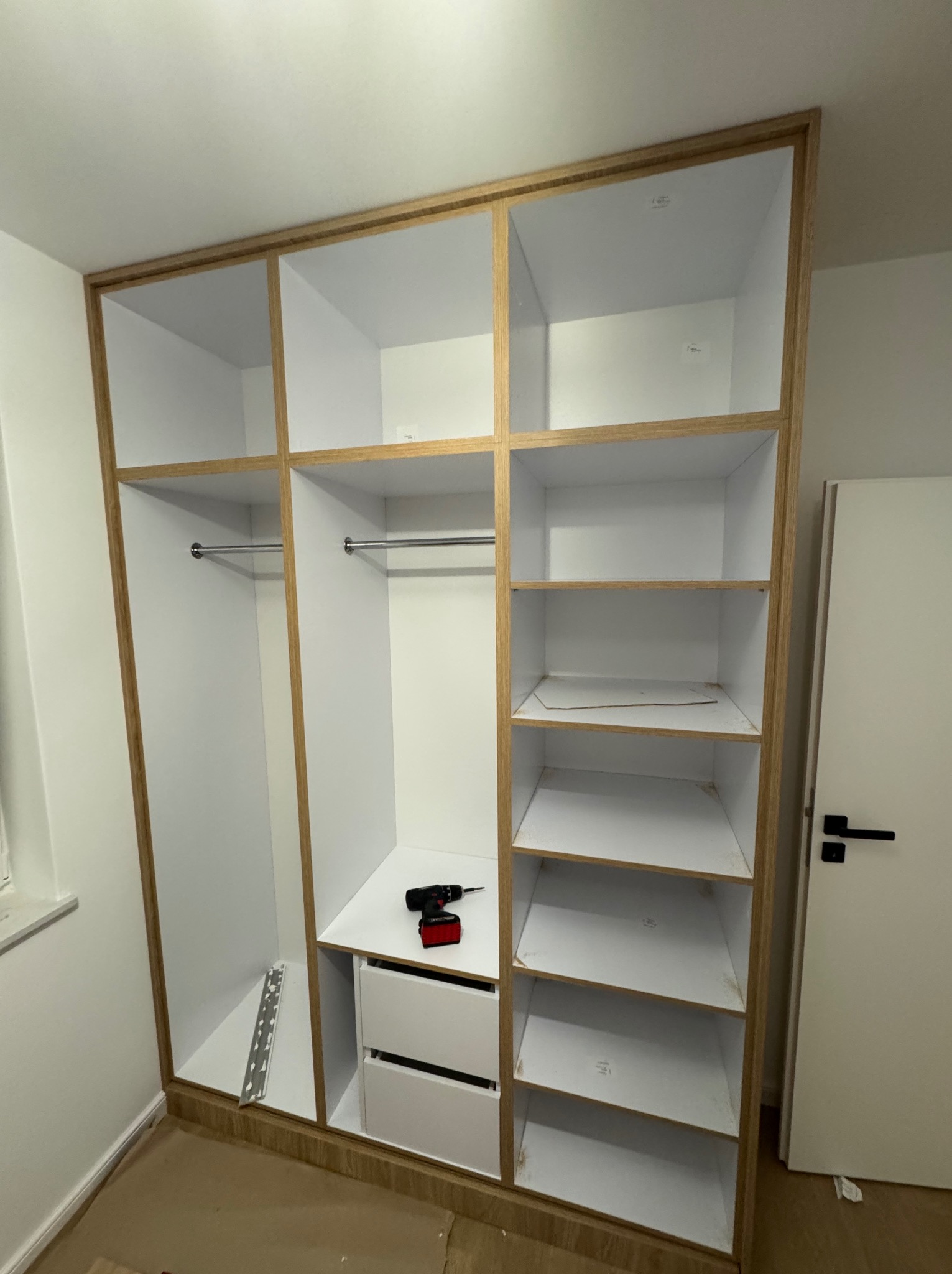 Wnętrze szafy na wymiar z białymi półkami i szufladami, obramowanie w kolorze jasnego drewna, w trakcie montażu widoczna wkrętarka i listwa montażowa.