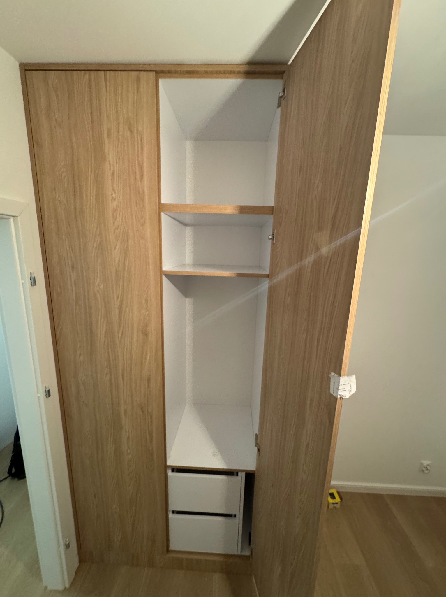 Wysoka szafa wnękowa z jasnego drewna z otwartymi drzwiami, widoczne półki i szuflady wewnątrz, białe wnętrze, minimalistyczny design.