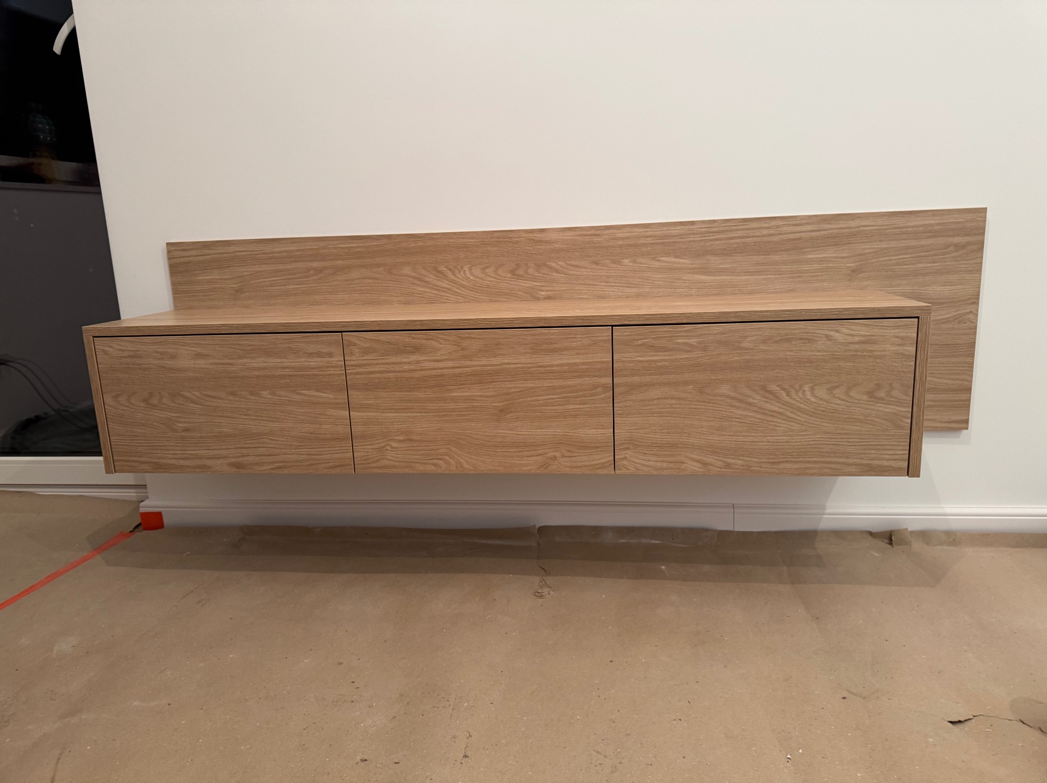 Wisząca szafka z trzema frontami z jasnego drewna z widocznym usłojeniem, zamontowana na białej ścianie, z fragmentem panelu ściennego z tego samego drewna. Podłoga zabezpieczona kartonem.