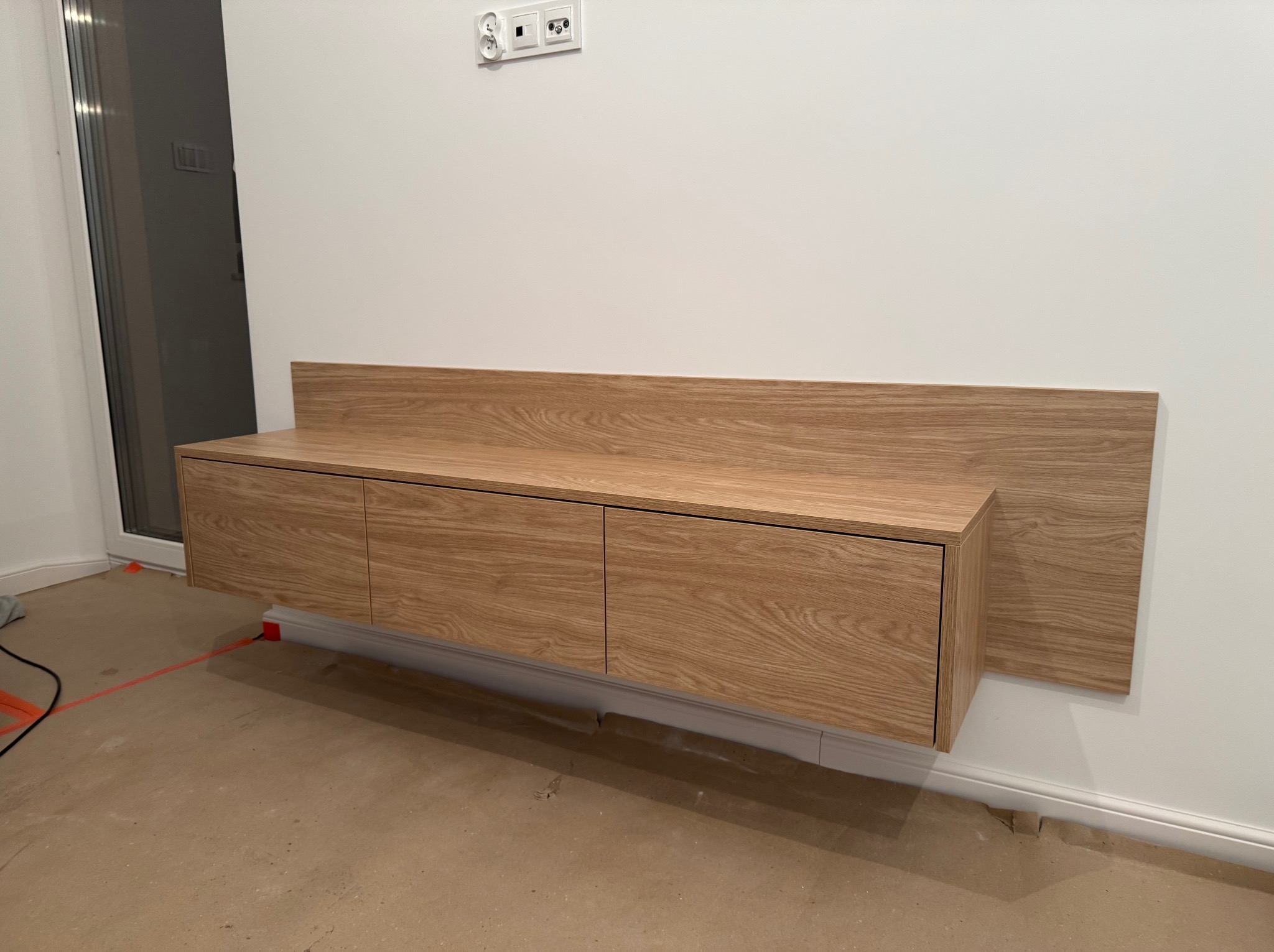 Wisząca szafka RTV z drewnianymi frontami i panelem na ścianie, widoczne gniazdka elektryczne i listwy przypodłogowe, podłoga zabezpieczona folią malarską.