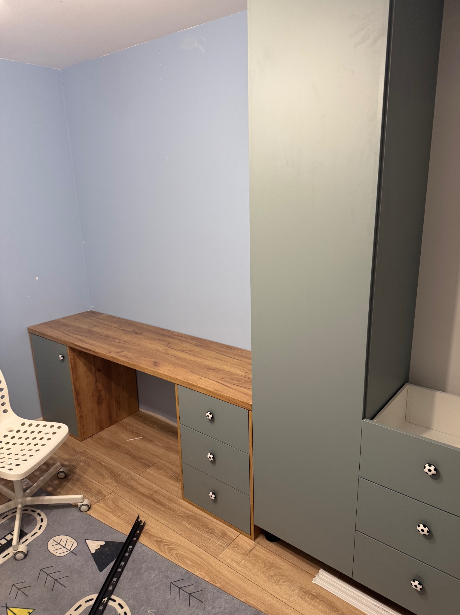 Szaro-drewniana zabudowa pokoju dziecięcego: szafa z szufladami i biurko z blatem imitującym drewno, uchwyty w kształcie piłek, białe krzesło na kółkach, dywan w szarościach z motywami dziecięcymi.