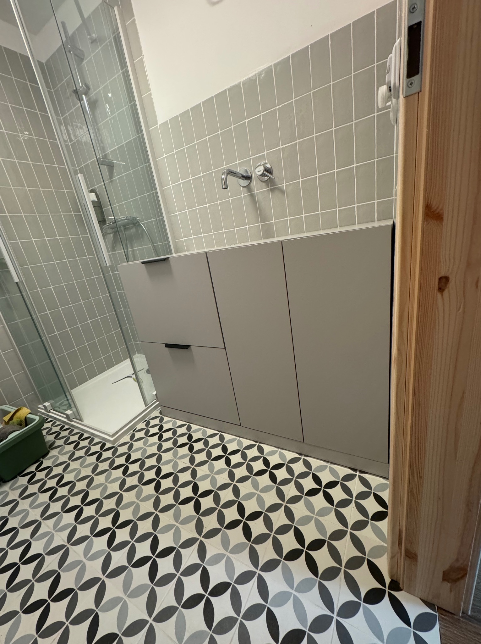 Szara szafka łazienkowa z czarnymi uchwytami pod umywalką, kabina prysznicowa ze szklanymi drzwiami i wzorzysta podłoga w łazience wykończonej szarymi płytkami.