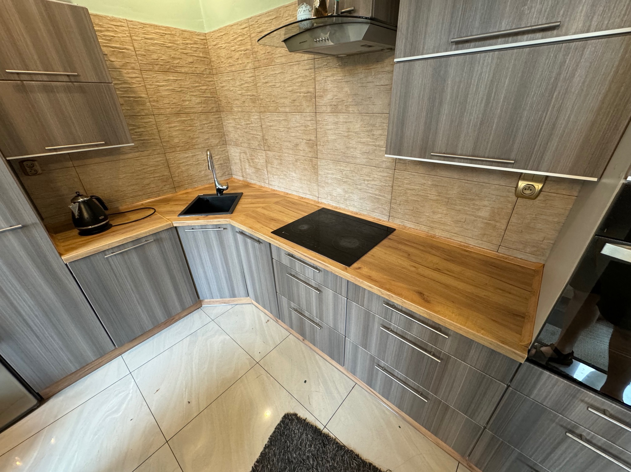 Nowoczesna kuchnia z szarymi szafkami i drewnianym blatem, czarna płyta indukcyjna, zlewozmywak i czajnik elektryczny na blacie, beżowe płytki na ścianie i jasne płytki na podłodze.