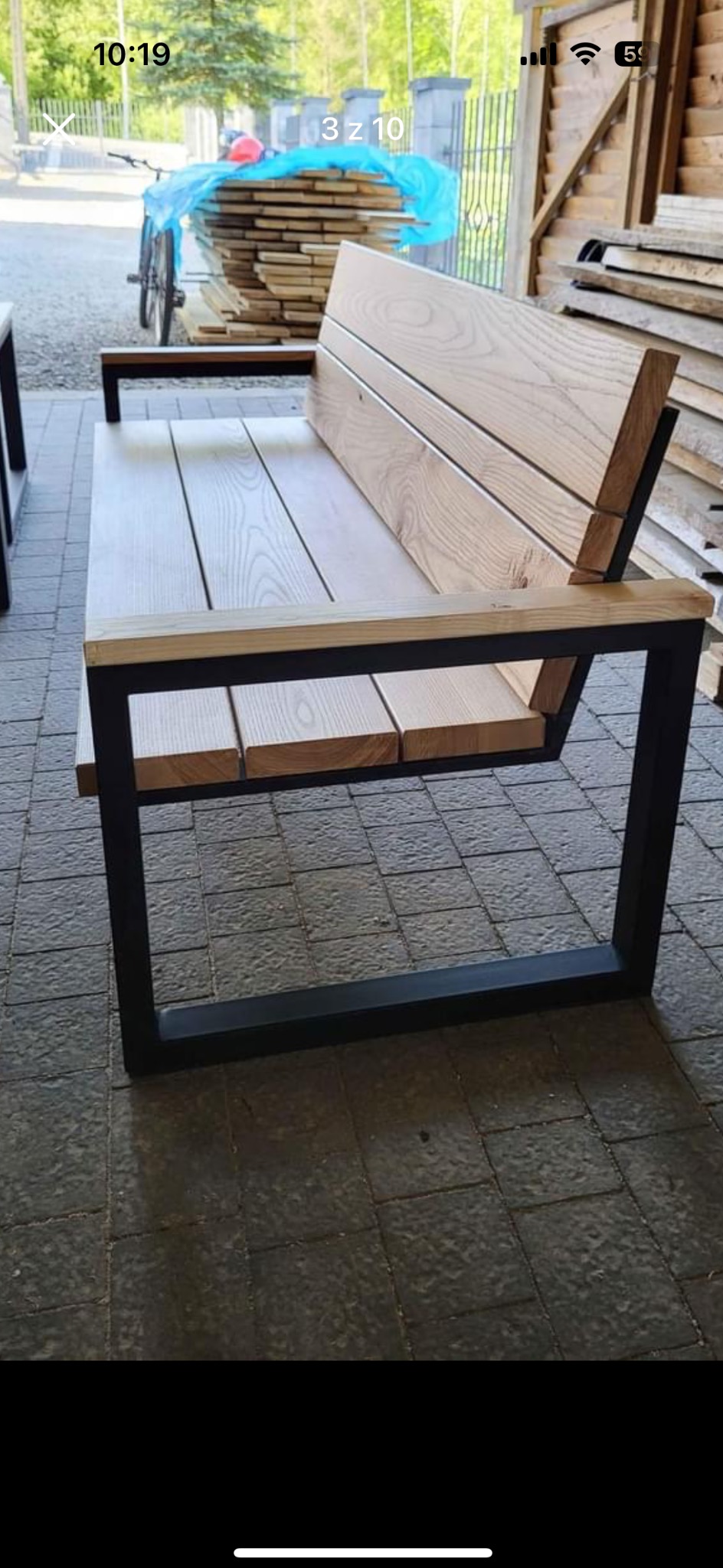 Drewniana ławka ogrodowa z metalową ramą, widoczna na tle ułożonego drewna i roweru, na szarym brukowanym patio.