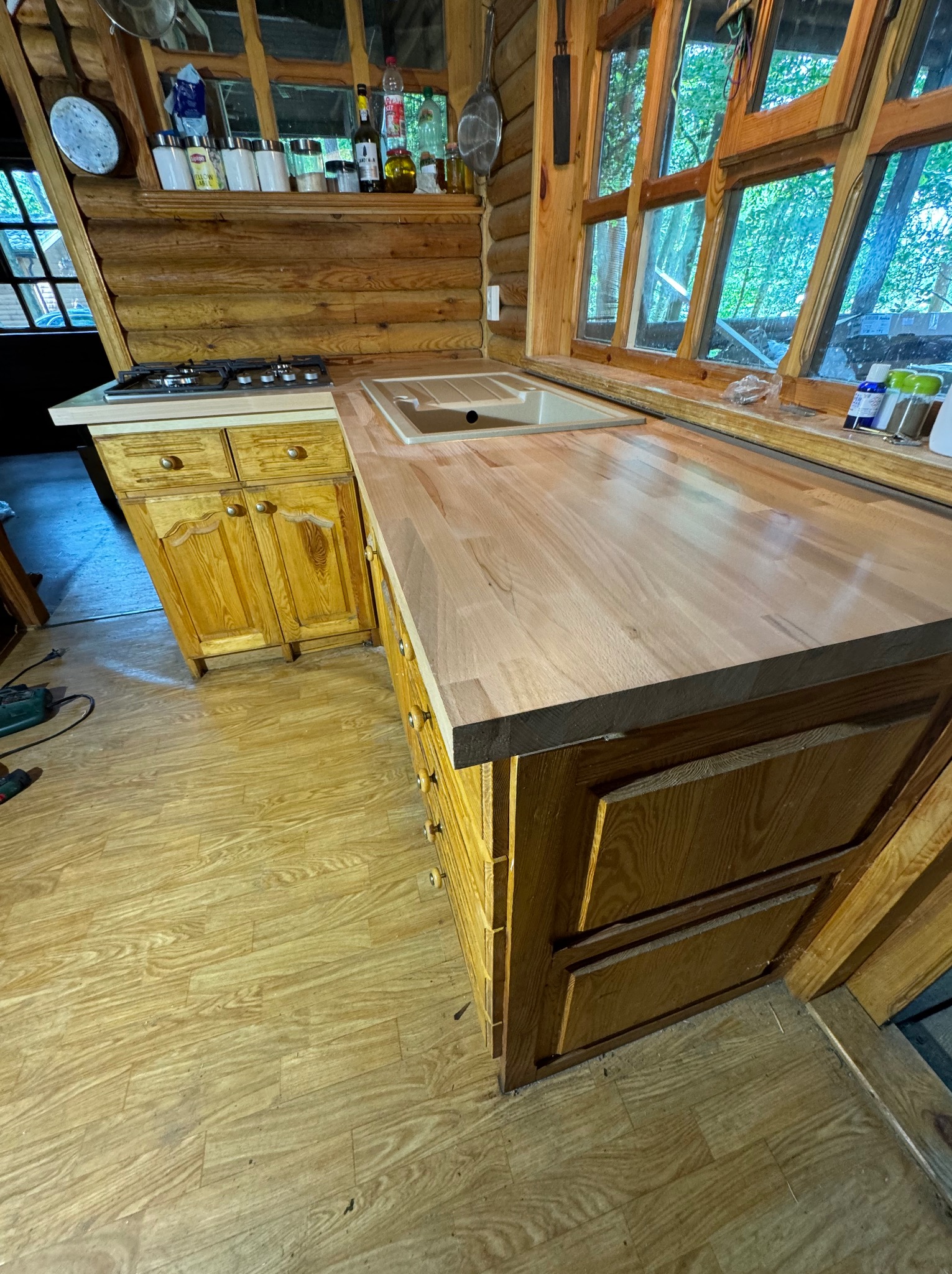 Surowy, jasny blat drewniany w kuchni z bali, widoczne słoje drewna, okno z widokiem na zieleń, szafki z jasnego drewna, podłoga imitująca drewno.