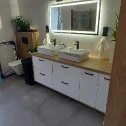 Nowoczesna łazienka z podwójną umywalką nablatową, oświetlonym lustrem, szarą podłogą i drewnianymi akcentami, widok z perspektywy wejścia.