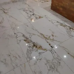 Błyszcząca, biała podłoga z płytek imitujących marmur, z widocznymi złotymi i szarymi żyłkami, świeżo położona w otwartej przestrzeni łączącej kuchnię i salon, z elementami ceglanej zabudowy...