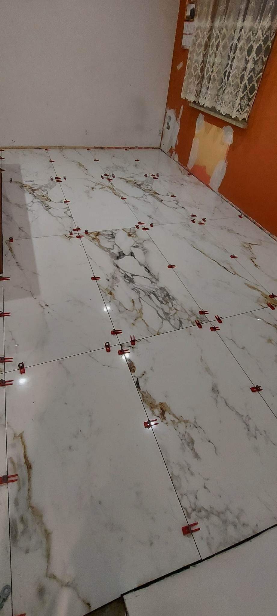 Ułożone płytki podłogowe imitujące marmur z czerwonymi klipsami poziomującymi, widoczne fragmenty przygotowanej ściany i okna z firanką.