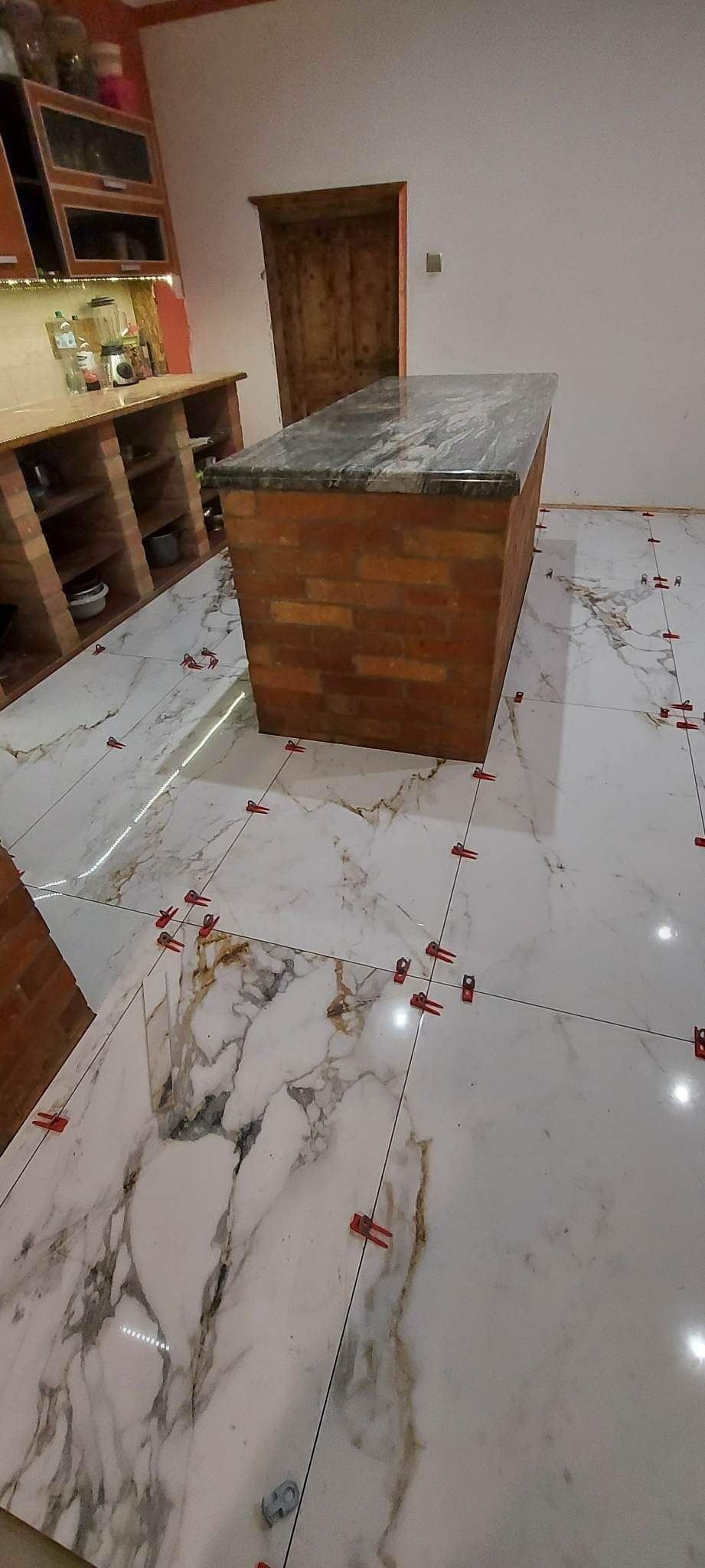 Białe płytki podłogowe imitujące marmur w trakcie układania, z czerwonymi krzyżykami dystansowymi, widoczny fragment wyspy kuchennej wykończonej cegłą i blatem z szarego kamienia.