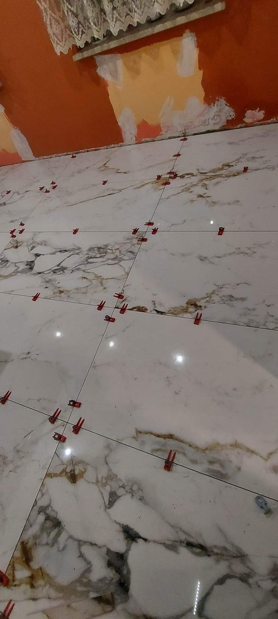 Podłoga wyłożona białymi płytkami imitującymi marmur z brązowymi żyłami, wyrównywana za pomocą czerwonych klipsów i czarnych krzyżyków dystansowych, ściana w tle przygotowana do malowania...