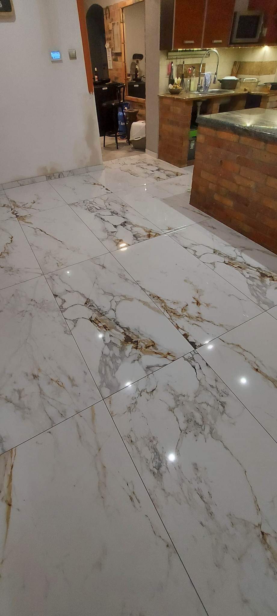 Błyszcząca, biała podłoga z płytek imitujących marmur, z widocznymi złotymi i szarymi żyłkami, świeżo położona w otwartej przestrzeni łączącej kuchnię i salon, z elementami ceglanej zabudowy...