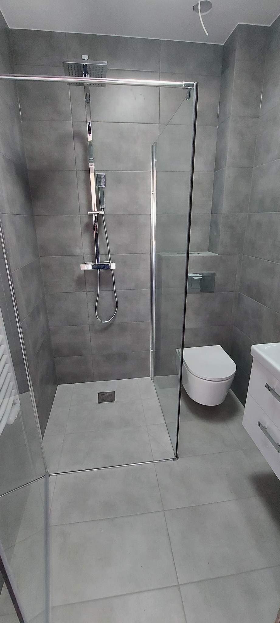 Nowoczesna łazienka z kabiną prysznicową typu walk-in, szarymi płytkami ściennymi i podłogowymi oraz minimalistyczną armaturą.