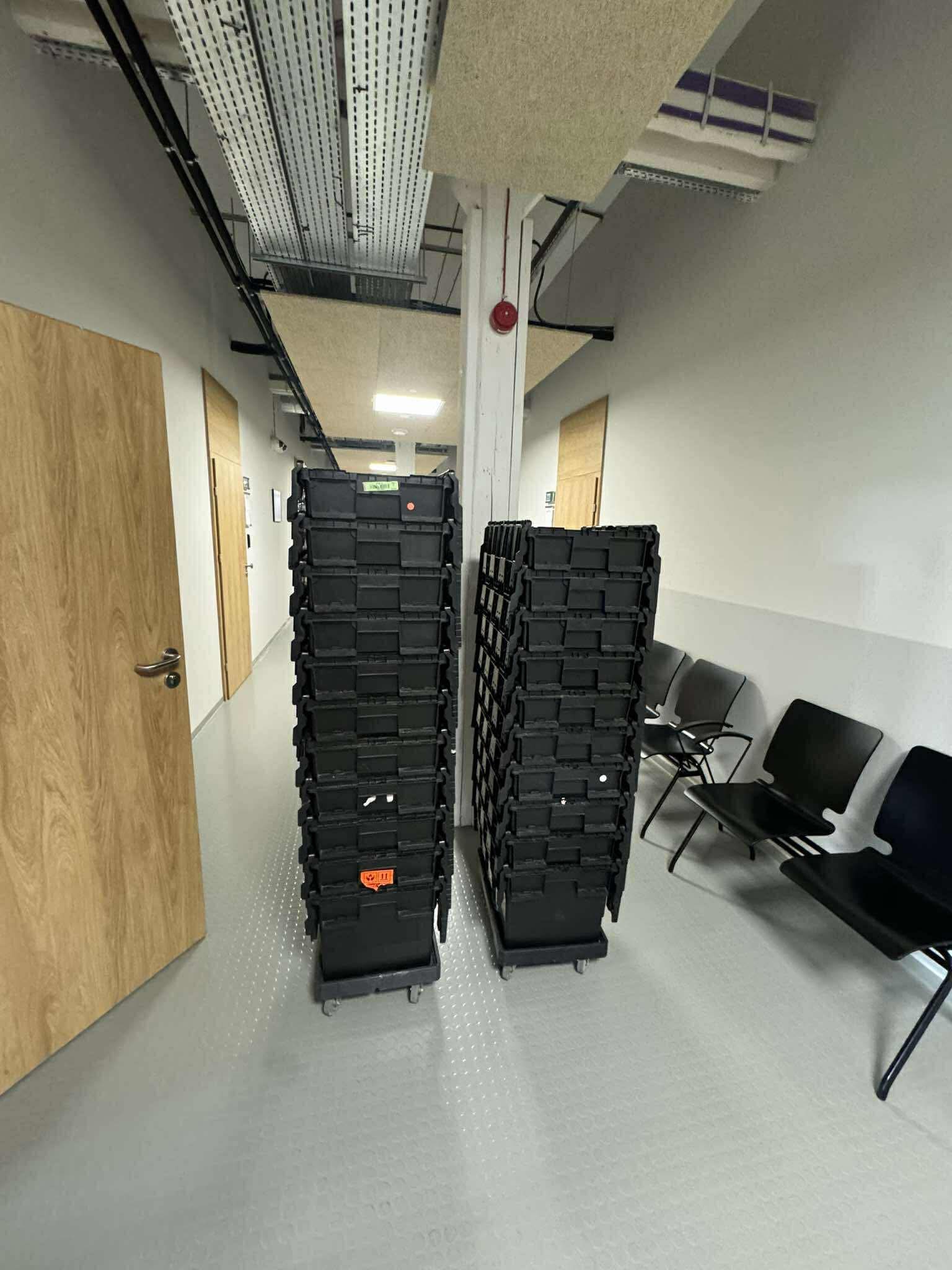 Dwa wysokie stosy czarnych, plastikowych pojemników na kółkach w korytarzu biurowym, gotowe do transportu. W tle krzesła i drzwi.