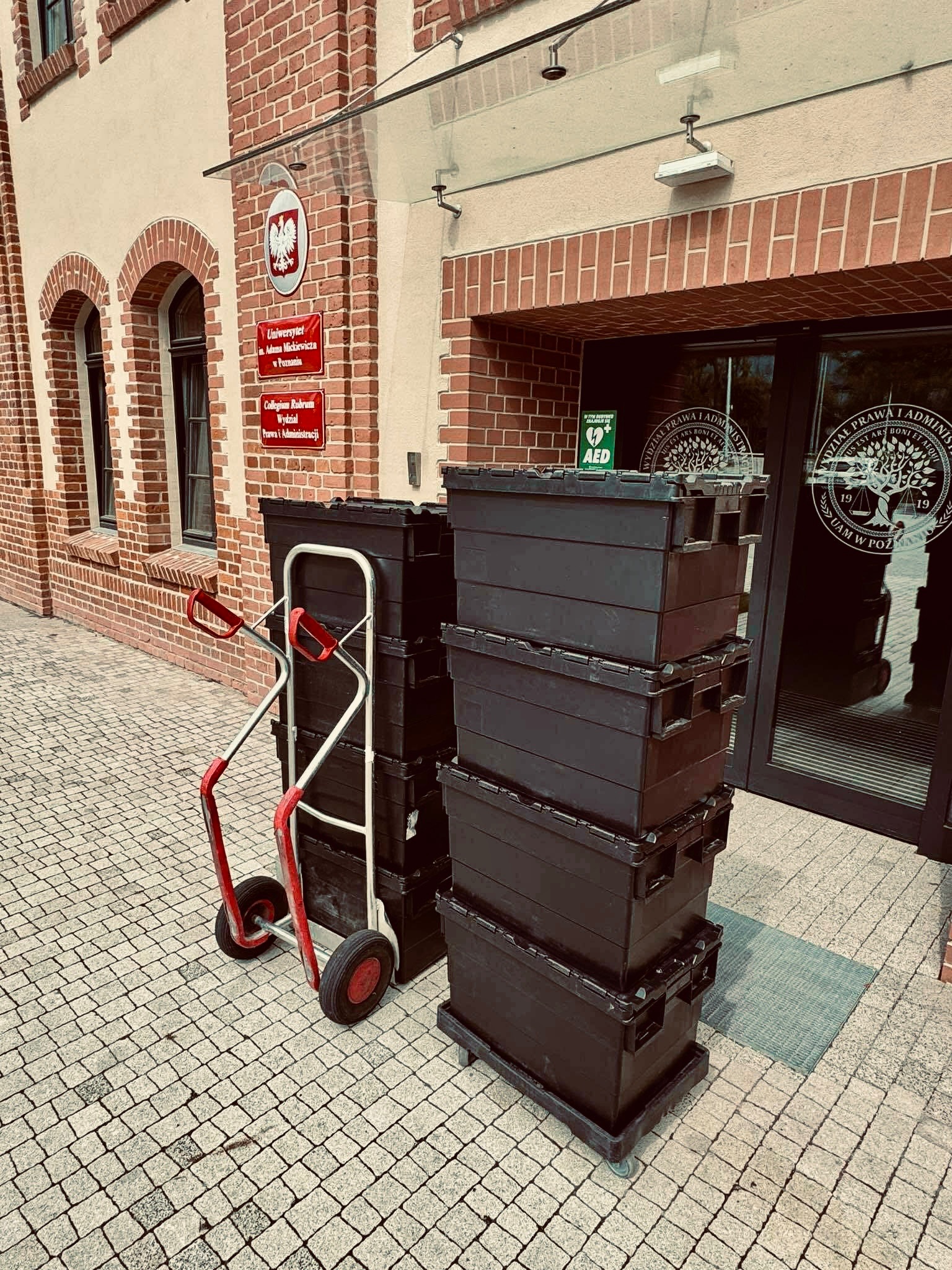 Stos czarnych skrzyń na wózku transportowym przed budynkiem Wydziału Prawa i Administracji UAM w Poznaniu. Przygotowania do przeprowadzki.