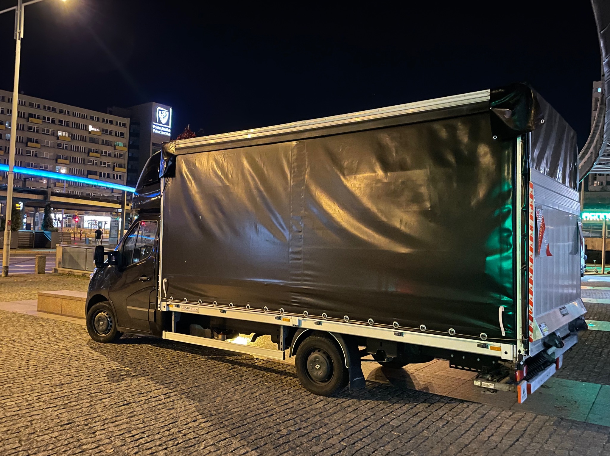 Czarny samochód dostawczy z plandeką zaparkowany nocą na brukowanej ulicy, w tle budynek Politechniki Wrocławskiej z podświetlonym logo.