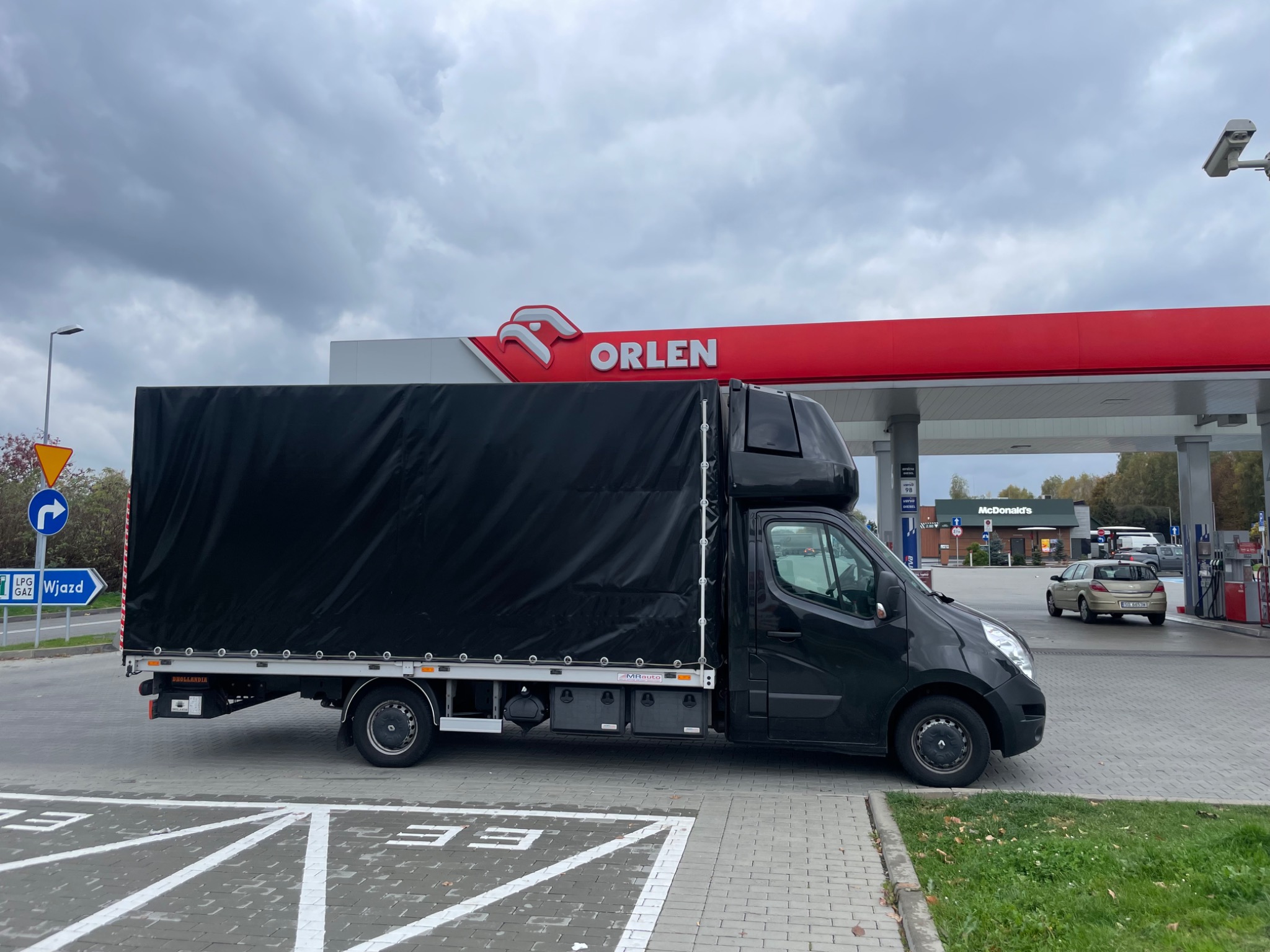 Czarny samochód dostawczy z plandeką na parkingu stacji Orlen, widoczny znak drogowy i budynek McDonald's w tle.