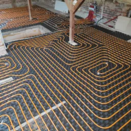 Pomarańczowe rury ogrzewania podłogowego ułożone na czarnych płytach montażowych w surowym wnętrzu poddasza z widoczną konstrukcją drewnianą i izolacją natryskową.