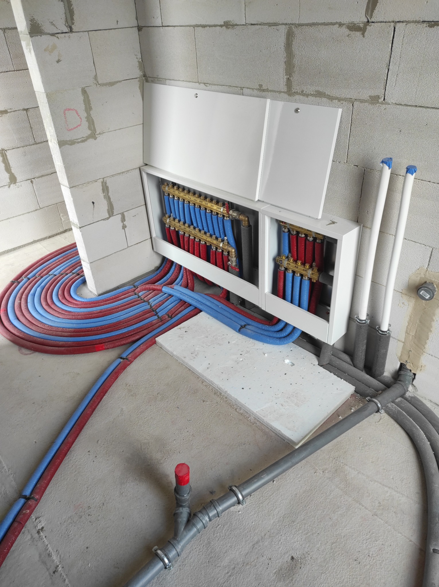 Rozdzielacz instalacji grzewczej podłogowej w białej szafce, z widocznymi pętlami rur w kolorach czerwonym i niebieskim na surowym betonowym podłożu i ścianach z betonu komórkowego.