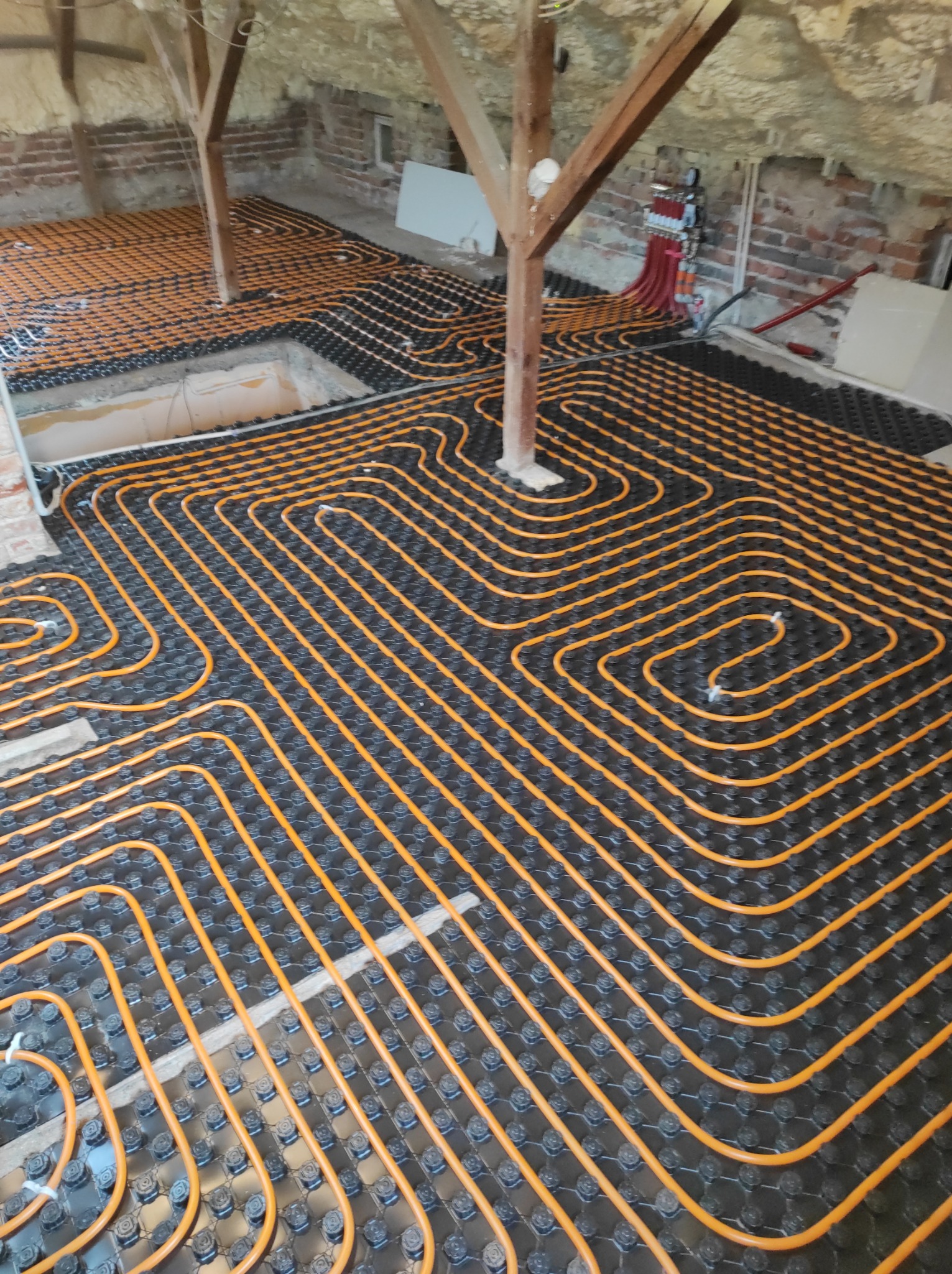 Pomarańczowe rury ogrzewania podłogowego ułożone na czarnych płytach montażowych w surowym wnętrzu poddasza z widoczną konstrukcją drewnianą i izolacją natryskową.