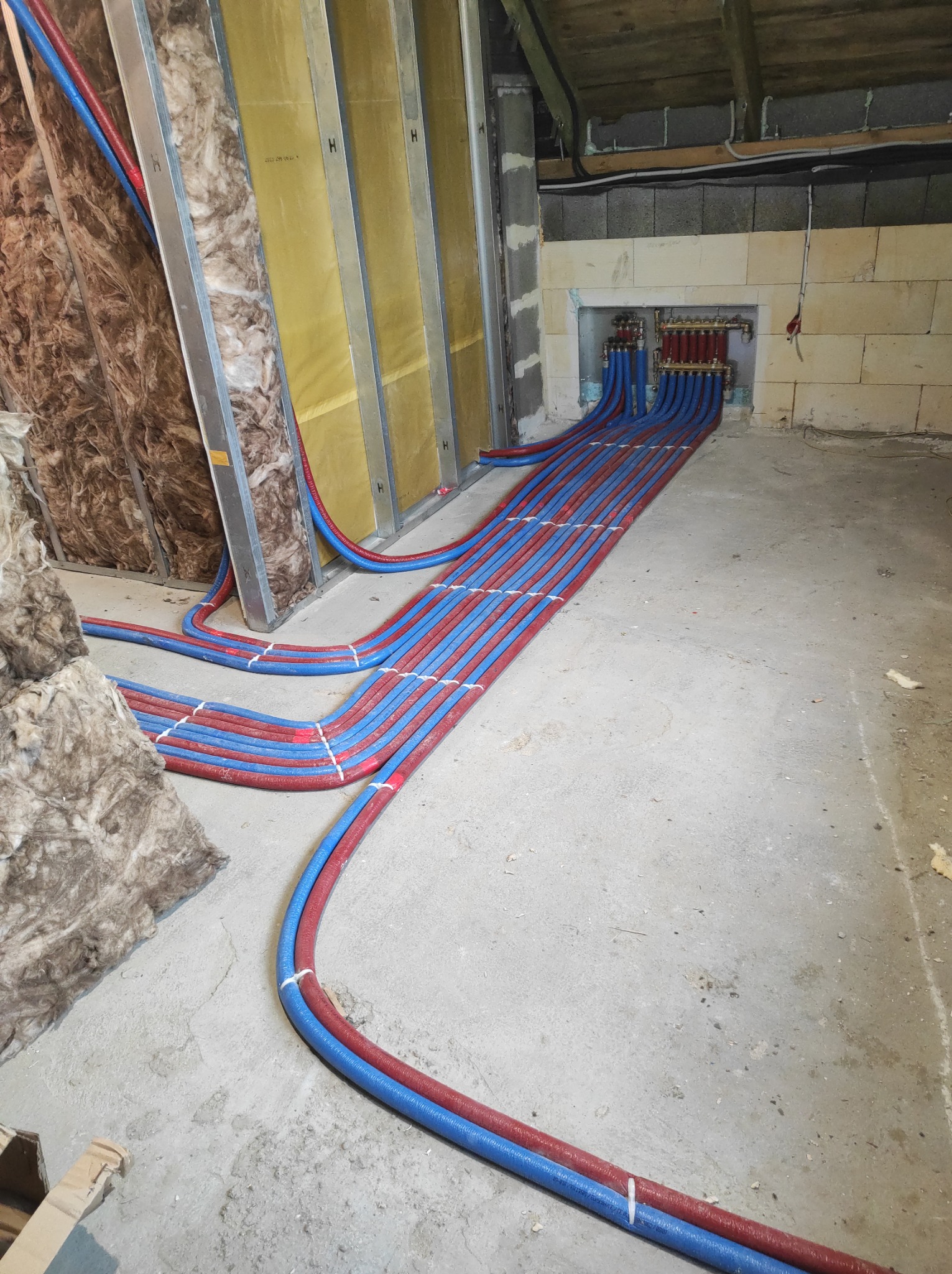 Rozdzielacz instalacji grzewczej z licznymi pętlami rur czerwonych i niebieskich, ułożonych równolegle na surowym betonowym podłożu w trakcie prac wykończeniowych, widoczne ocieplone ściany i sufit.