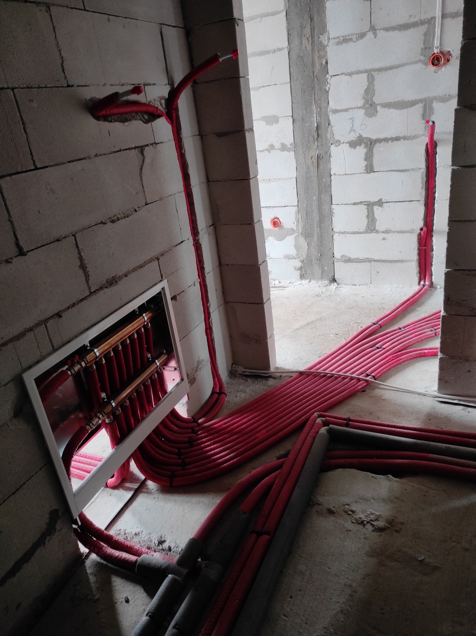 Rozdzielacz ogrzewania podłogowego z czerwonymi rurkami rozprowadzonymi po surowym wnętrzu w trakcie instalacji, widoczne bloczki betonowe i wylewka w tle.