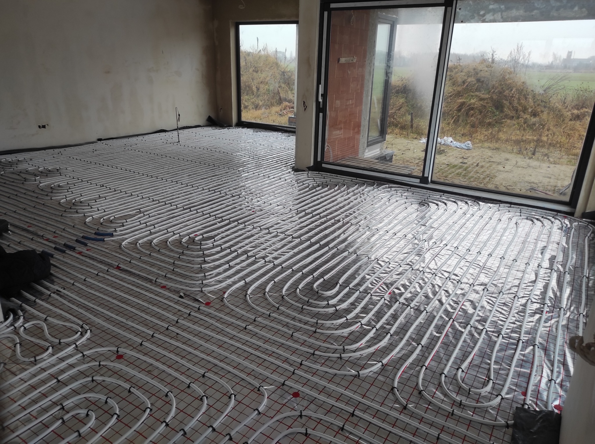 Białe rury systemu grzewczego ułożone na folii z czerwoną siatką w surowym wnętrzu z dużymi oknami.