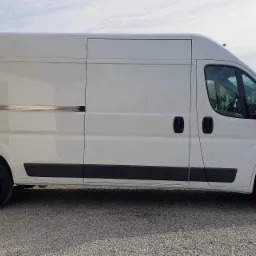 Biały samochód dostawczy Citroen Jumper na żwirowym podłożu, widok z boku, zarys zabudowań w tle.