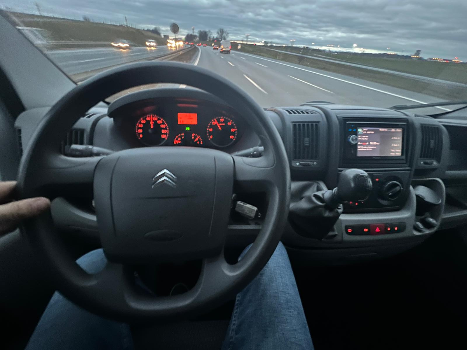 Widok z wnętrza kabiny Citroena Jumper podczas jazdy autostradą, deska rozdzielcza z widocznymi zegarami i radiem, kierowca w jeansach.