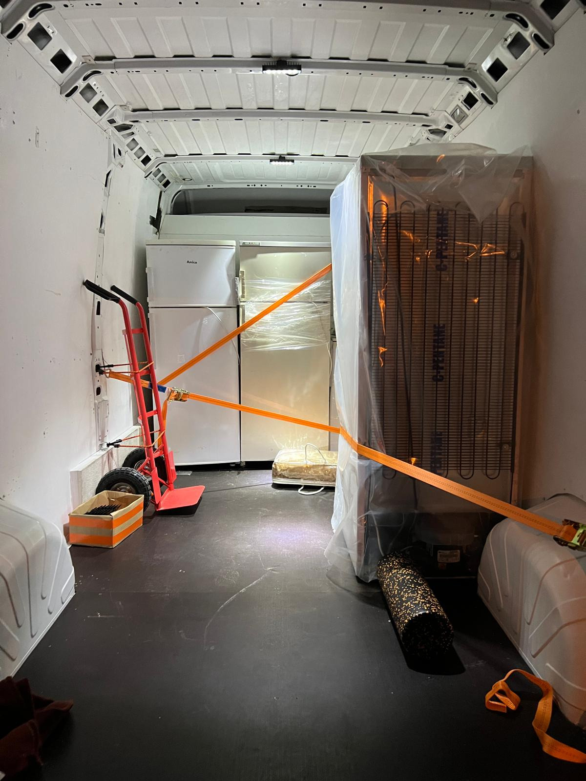 Wnętrze furgonetki z zabezpieczonymi lodówkami marki Amica i owiniętą folią lodówką transportowaną za pomocą wózka transportowego z pomarańczowymi pasami mocującymi.