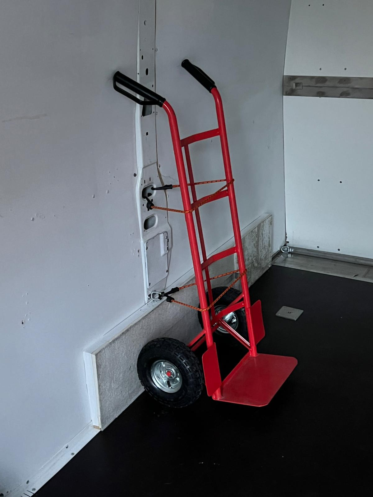 Wnętrze furgonetki z czerwoną platformówką do transportu, zabezpieczoną elastyczną liną, przy ścianie z widocznymi mocowaniami.