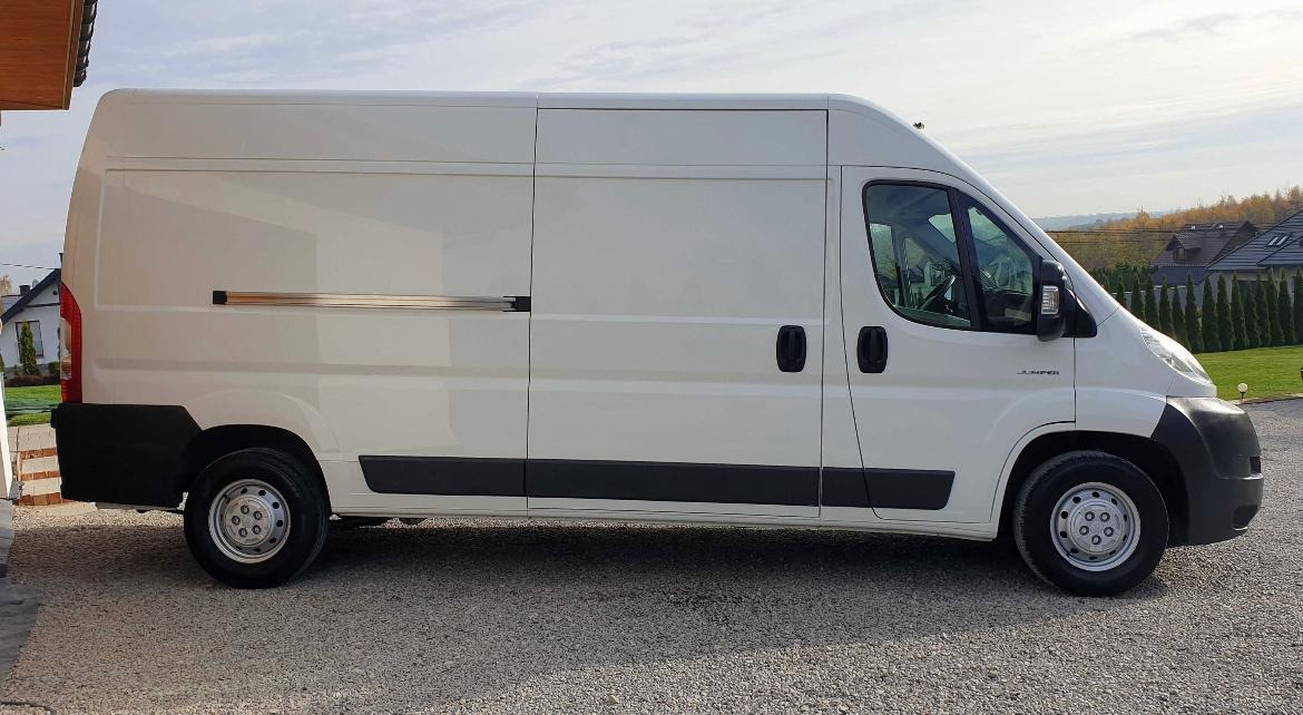 Biały samochód dostawczy Citroen Jumper na żwirowym podłożu, widok z boku, zarys zabudowań w tle.