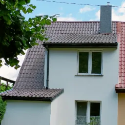 Dwupiętrowy dom z białą elewacją, brązowym dachem z dachówki i szarym kominem; widoczne okna z firankami i żółta skrzynka pocztowa.