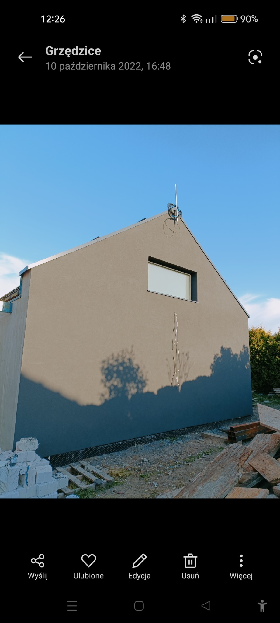 Świeżo otynkowana elewacja domu jednorodzinnego w odcieniach szarości, z widocznym fragmentem okna i wystającą anteną na dachu, w tle błękitne niebo i cień drzewa na ścianie, na ziemi materiały...