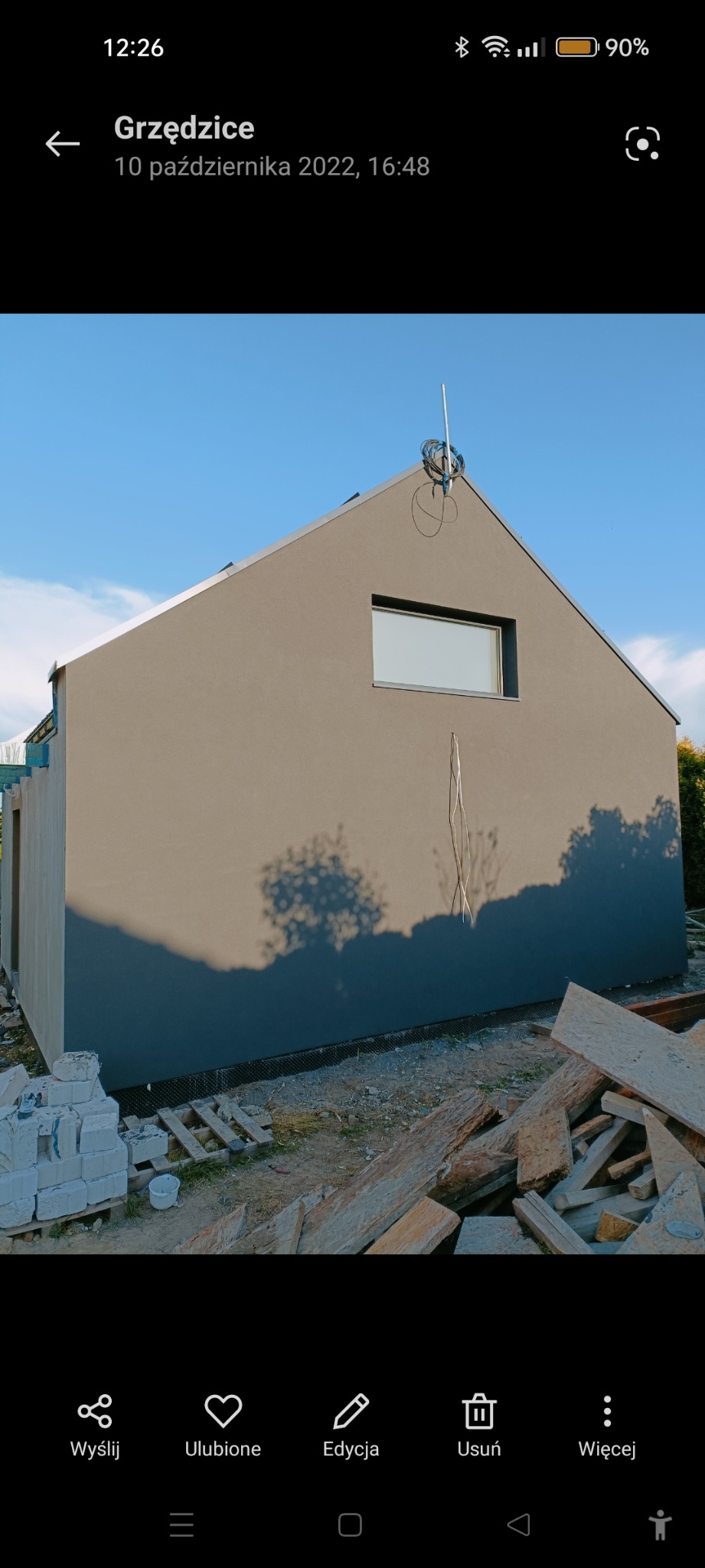 Ściana domu z szarą elewacją i pojedynczym oknem, widoczne materiały budowlane i cień drzewa, na dachu antena.