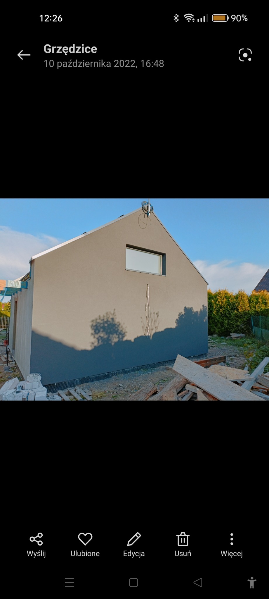 Świeżo otynkowany budynek gospodarczy z małym oknem, widoczne pozostałości materiałów budowlanych i cień drzewa na elewacji.