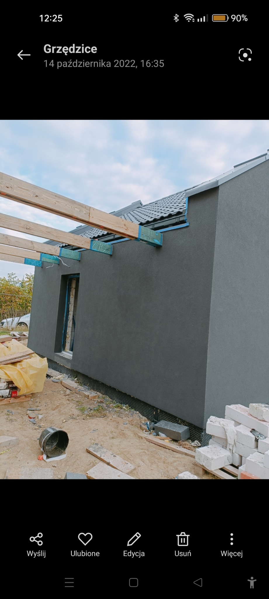 Szara elewacja budynku w trakcie prac wykończeniowych, widoczne drewniane elementy konstrukcji dachu zabezpieczone niebieską taśmą, oraz stos białych cegieł na ziemi.