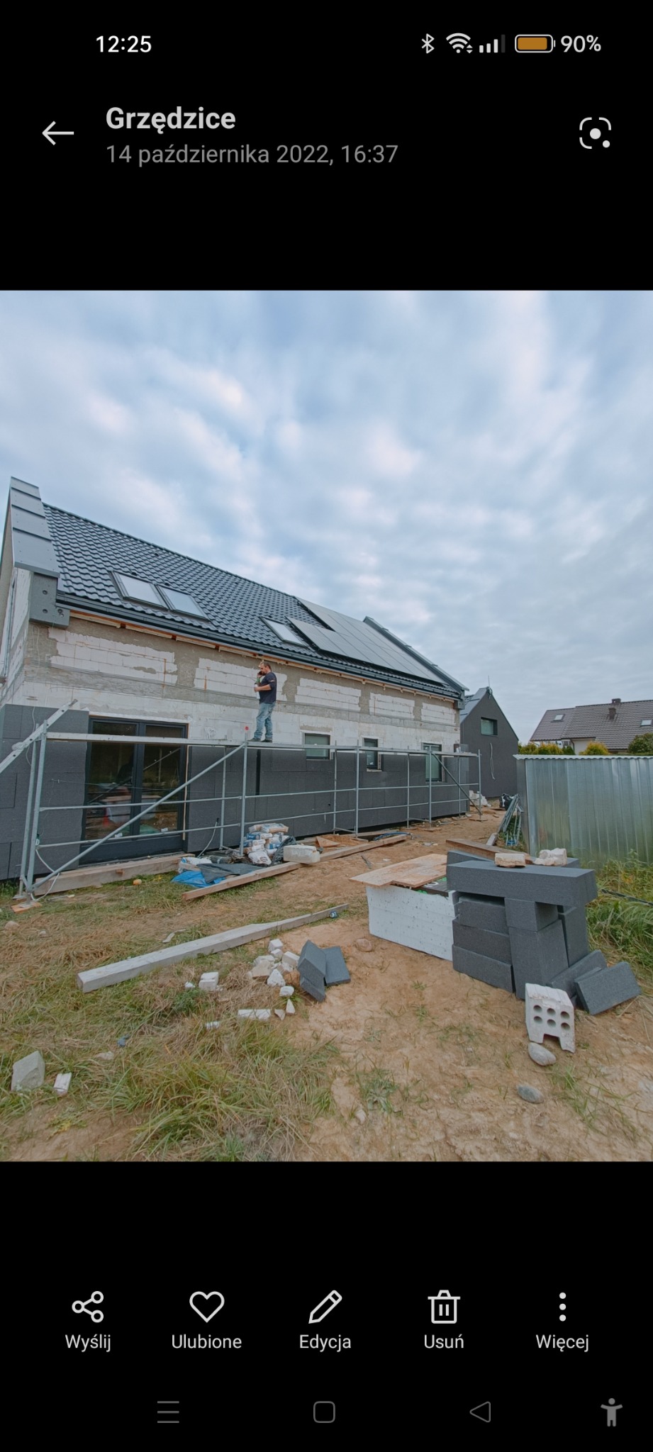 Budynek w trakcie prac wykończeniowych elewacji, widoczne rusztowania, płyty izolacyjne oraz pracownik na rusztowaniu. Na dachu zamontowane panele słoneczne.