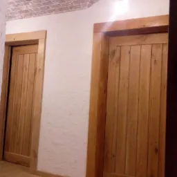 Dwa drewniane skrzydła drzwiowe z ościeżnicami w jasnym odcieniu drewna, widoczne na tle białej ściany o chropowatej fakturze i sklepienia z cegły, oświetlone pojedynczą żarówką.