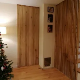 Drewniane listwy pionowe tworzące ażurową ściankę działową między pokojami, z widocznymi schodami i choinką w tle, oświetlenie ciepłe, parkiet na podłodze.