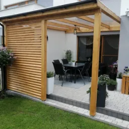 Zadaszony taras z drewnianą konstrukcją i lamelową osłoną, wyposażony w czarny stół z krzesłami, dekorowany roślinami w donicach, z widokiem na trawnik.